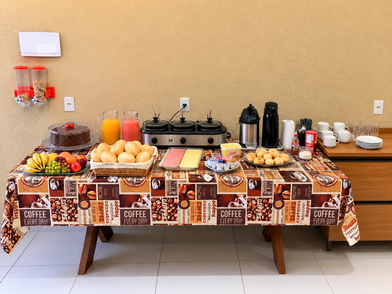 Mesa de café da manhã do Hotel Ideal 2 com pães, frutas, bolo, sucos e café
